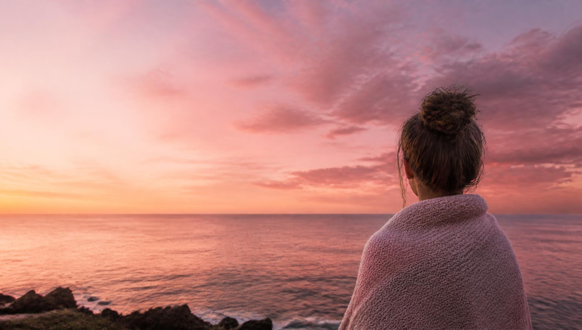 A woman wrapped in a soft blanket looking out over the sea at sunset
